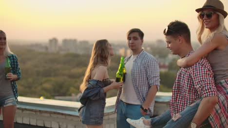Loving-couple-moves-in-a-dance-at-a-party-with-friends-who-drink-beer.-A-girl-sits-on-her-boyfriend's-back-they-are-fooling-around-on-the-roof.